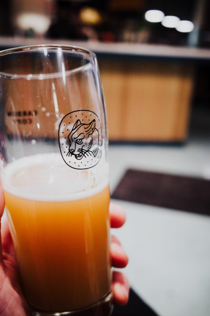 Service-1-min (1) Close-up image of a hand holding a craft beer glass with a unique logo in a cozy bar setting.